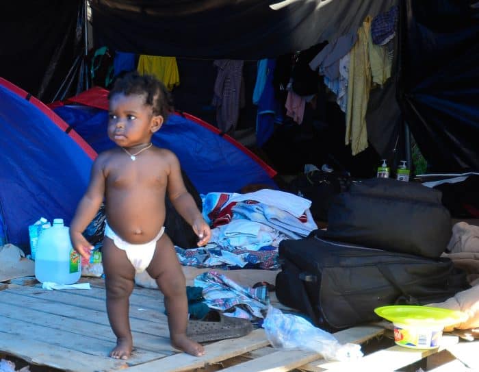 A baby in a diaper stands among tents and bags at the Paso Canoas border camp.