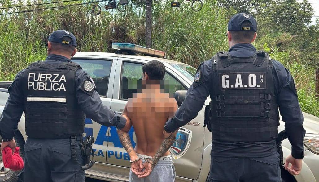 Costa Rican Fuerza Pública officers detain a handcuffed man beside a police vehicle in San Carlos.
